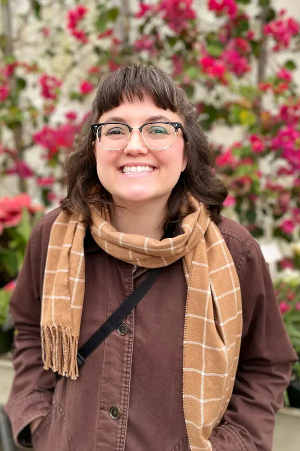 White woman with shoulder length brown hair and glasses wear a brown coat and tan scarf, standing against a background of red flowers.nt background. 