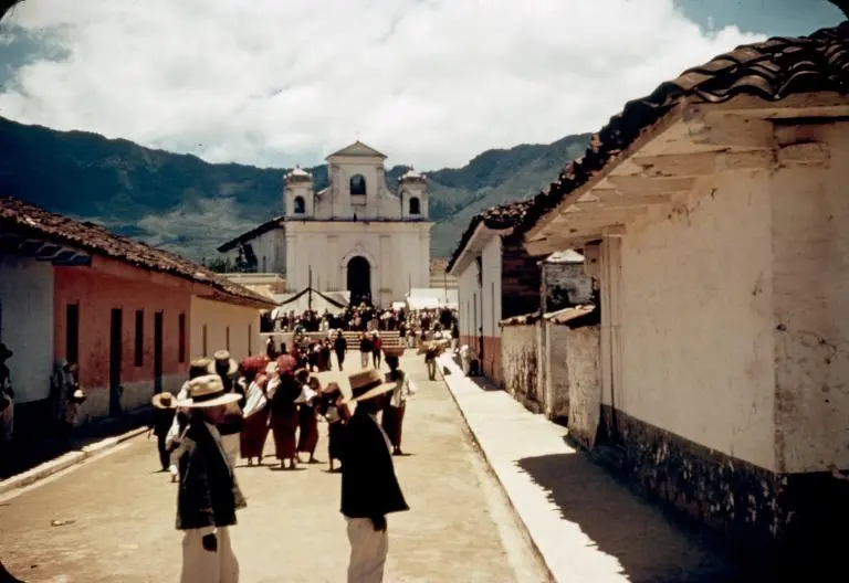 A 4 story white church and green mountains stand behind a distant, people-filled town square flanked by one story buildings and people milling in the street in the foreground.