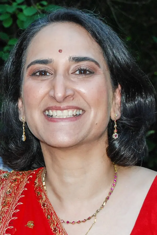Head and shoulders image of a dark-haired woman with an olive complexion, wearing a red sari and smiling broadly