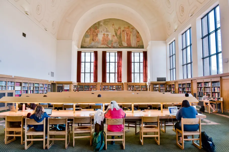 Wide view of the iconic reading room with large windows, students at long tables, and a mural in the distance.